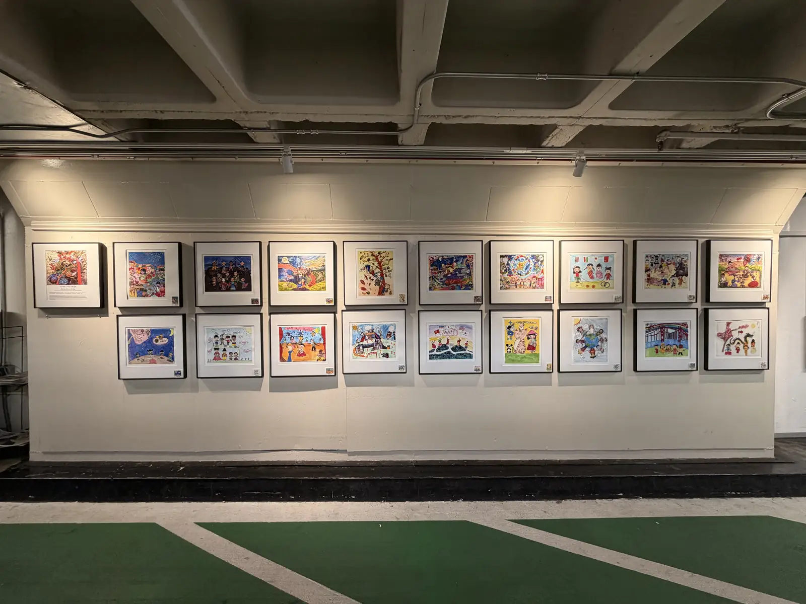 Two rows of framed children's paintings from the I Love Chinatown Children's Art Contest on the exhibition wall inside the garage